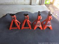 Four red metal car jack stands arranged on a black tarp surface in a garage or outdoor sheltered area.