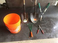 Photo showing assortment of garden tools including orange bucket, two shovels, hedge shears, garden rake, and a pair of hand pruners on black surface.