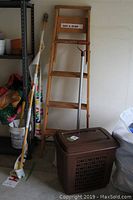 Full lot shot showing the wooden ladder, metal shovel leaning on the ladder, and brown plastic laundry hamper in front of the ladder.