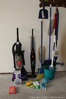 Photo of Bissell Powerforce Helix vacuum, Oreck Dual Power vacuum, mop with bucket, broom, duster, umbrella, Bona hardwood floor cleaner, and assorted cleaning supplies arranged against wall with hooks.