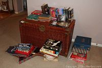 Top view photo showing a wicker trunk topped with assorted books, board games, VHS tapes, cassettes, and CDs. Additional items arranged around the trunk including board games and books.