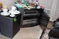 Side view of media cabinet showing Ikko VHS player on upper shelf, Panasonic cassette receiver on lower shelf, and one black speaker next to the cabinet.