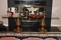 Full view of glass coffee table with two ornate bronze/gold colored decorative bases, rectangular clear glass top, set against a fireplace background with household items on top.