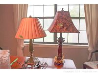 Two table lamps positioned on a wooden surface in front of window with curtains. One has tan pleated shade, the other a patterned square shade with floral design.