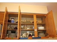 Photo showing wooden kitchen cabinets with glass doors containing stacked white dishes, clear glass bowls, and small serving bowls including a sugar bowl and teapot.