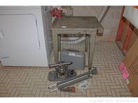 Essco surveying tool mounted on tripod with case, metal table with wood top, vise attached, all on basement floor next to dryer.