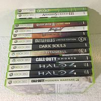 Photo showing 13 Xbox 360 game cases stacked horizontally, titles visible on the spine: Xbox Live Arcade, Assassin's Creed III, Guitar Hero Legends of Rock, Aerosmith, Dark Void, Battlefield 3 Limited Edition, Dark Souls, Titanfall, Call of Duty Ghosts, Halo 4 (two copies), Call of Duty Modern Warfare 3.