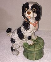 Porcelain figurine of a King Charles Spaniel dog with black and white markings sitting with one paw on a green tufted stool.