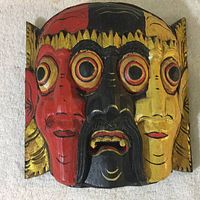 Hand-carved and painted three-faced wooden mask in red, black, and yellow hues with detailed facial features and large circular eyes.