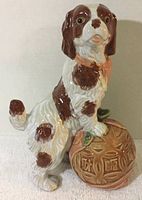 Side view of porcelain King Charles Spaniel figurine with white and brown colors standing on a Chinese ball with carved patterns.