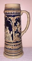 Front side of the stein showing a detailed scene of a woman and man in traditional attire with ornamental scrollwork and grapevine decorations.