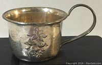 View of the vintage sterling silver baby cup showing the floral engraving on the side and handle, with visible dings and surface wear.