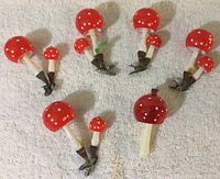 Six glass mushroom ornaments with orange caps and white polka dots with metal clip bases on a white cloth background showing various sizes and some rust on clips and paint loss on stems.