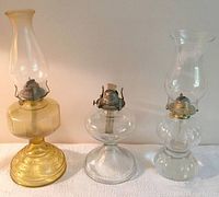 Lot of three vintage glass oil lamps on white surface against cream background. Amber colored lamp on left, clear glass middle lamp without shade, clear glass right lamp with chimney.