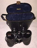 Manon 10x50 binoculars standing in front of open black leather case with blue velvet interior and Manon logo inside lid.