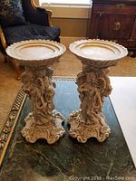 Photo showing pair of beige molded resin angel candle holders from front, with detailed angel figures holding books and ornate bases.