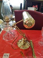 Photo showing the brass adjustable desk lamp with cylindrical shade and round base on a red surface near the glass oil lamp.