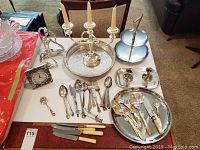 Photo of table with vintage cutlery and serving pieces including butter knives, forks, spoons, a 3-branch candle holder, small clock, and tiers serving tray.