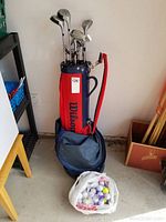 Full view of golf bag with clubs and bag of golf balls on concrete floor beside shelf and boxes.