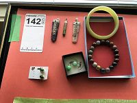 Collection of semi precious jewelry arranged for display including jade bangle, bead bracelet, pendants, and earrings on orange surface