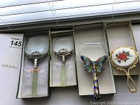 Four magnifying glasses displayed in green-lined boxes showing different decorative handles including jade-style with silver metal and green stones, cloisonné butterfly, and floral painted rose handle.