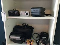 Photo of shelf with Canon 8mm video camera, Canon Sure Shot Telemax, Sony Cyber-shot, Polaroid camera, camera case, film rolls and magnifying glass.