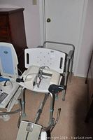 Photo showing the white plastic shower chair with backrest, armrests, and metal frame supports along with parts of the recliner and footrests.
