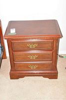 Solid wood bedside table with three drawers and brass handles in a carpeted room.