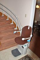 Electric stairlift chair with brown cushion and white armrests, positioned at the base of a carpeted curved staircase with white metal rail attached.