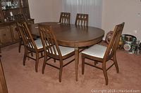 View of oval solid wood dining table with leaf surrounded by six matching chairs with white upholstered seats, set on carpeted floor.