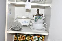 Close-up of middle shelves showing Corningware casserole dish, glass sugar bowl, teapot with cup/saucer, and decorative tile trivet on wrought iron holder.
