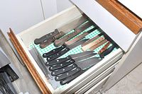 Drawer with assorted kitchen knives having black and wooden handles laid on drawer liner.