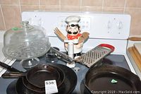 Top view of stove with cookware including two cast iron pans, metal grater, ceramic utensil holder shaped like chef, wooden utensils, and glass dome on cake stand.