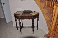 Solid wood table with turned spindle legs positioned against a wall beneath staircase, with framed bird prints and plaques on top.