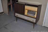 Side and front view of vintage Barbizon stereo console in a basement. Wood veneer finish, flip-top lid open showing record player inside, radio tuner with analog dials behind glass panel.