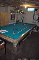 Full view of pool table in basement setting showing woodgrain frame, green felt surface, two cues and burner light fixture overhead