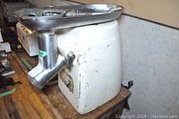 Side view of the white metal electric meat grinder with stainless steel grinder attachment and tray placed on wooden tabletop.