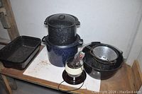 Photo showing black and blue enamel speckled stockpots, metal colander, round black bowl, and electric coffee percolator on shelf.