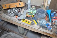 Wide view of a workshop bench with assorted hand tools spread across the surface, showing rulers, pliers, knives, and a wooden case.