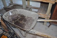 Metal wheelbarrow with rusted interior and wooden handles, placed indoors near wooden shelving.