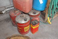 Photo showing assorted vintage red metal gas cans and blue water jugs near a wall with plumbing and green garden hose.