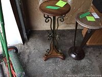 Photo showing two small accent tables. One table has a round wooden top with a decorative metal scrollwork base and wooden feet. The other table has a round stone top with a simple metal base.