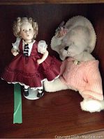 Shirley Temple figurine standing next to the stuffed bear on a wooden shelf, showing full figure and bear's bonnet and sweater.