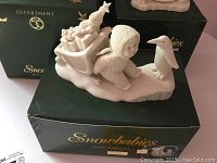 First detailed photo showing one of the Snow Babies figures with a child in a textured winter outfit beside a penguin on a sled, resting on original green box with gold Department 56 logo.