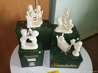 Photo of six Snowbabies figurines placed on three dark green Snowbabies branded boxes, showing varied poses including children sitting and interacting with objects.