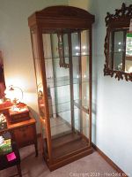 Full view of the tall wood veneer curio cabinet with glass front and sides, standing in a corner near other furniture.