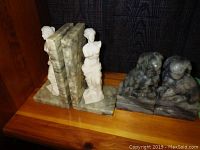 Two pairs of stone bookends displayed side by side: classical female statues on the left and Foo Dogs on the right on a wooden shelf against dark background.
