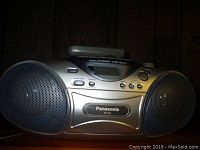 Front view of the silver Panasonic RX-D20 portable stereo system on a wooden surface, showing the digital tuner, control buttons, and left speaker.