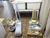 Electrolux upright vacuum cleaner with accessories, Aerus vacuum, and Dirt Devil handheld vacuum shown together on floor by laundry sink.