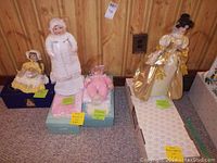 Four dolls on display: Three children/infant style dolls on boxes, one dressed in white winter attire, one baby doll in pink, one child in yellow bonnet and dress; one large doll in elaborate gold and cream gown standing next to them.